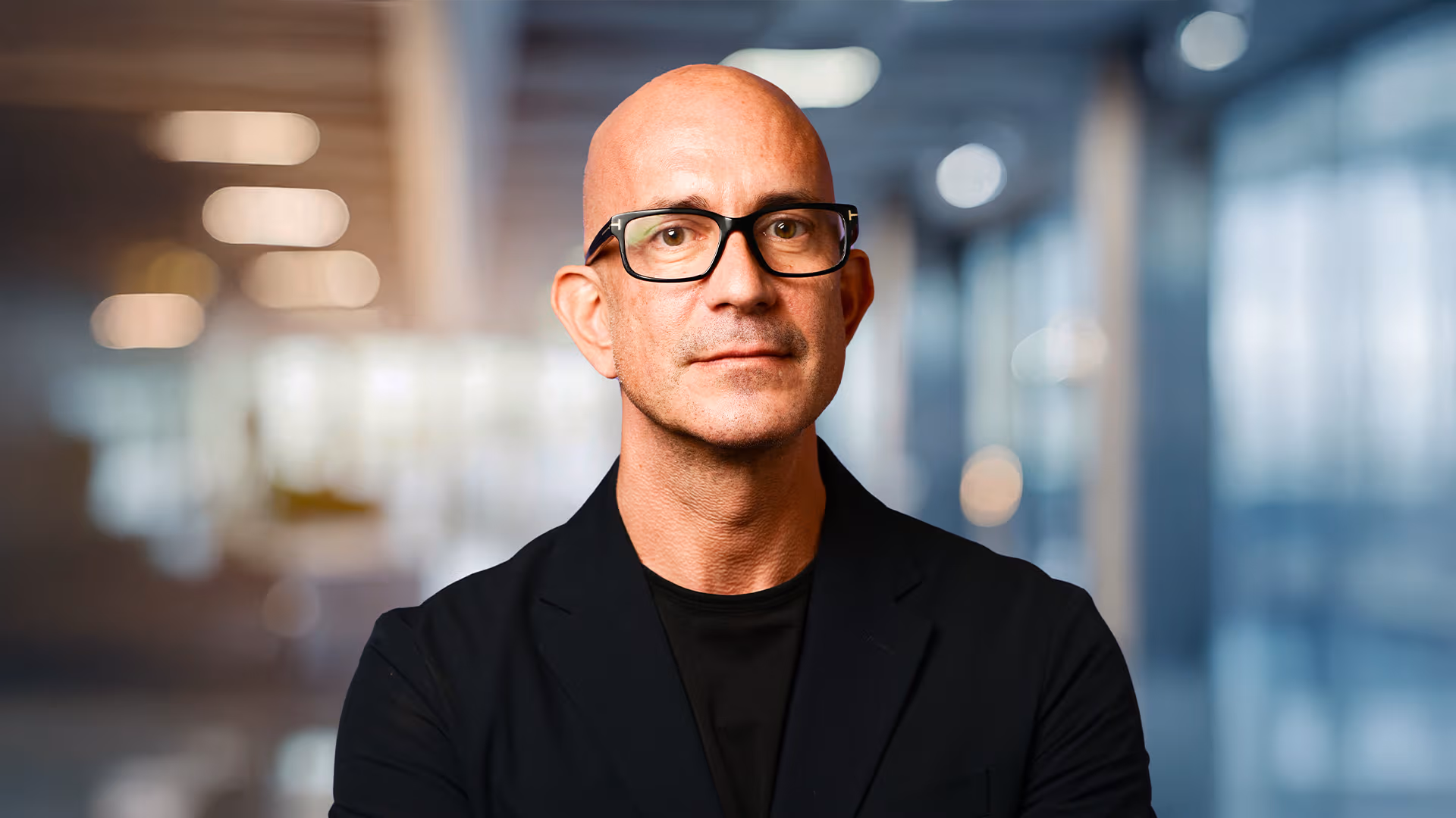 Bald man wearing black glasses and a black blazer, standing in a modern office with blurred background.